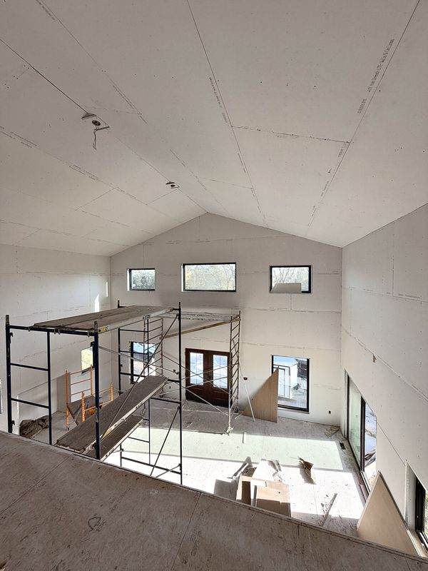 Vaulted great room with fresh drywall, tall windows and scaffolding, viewed from the loft